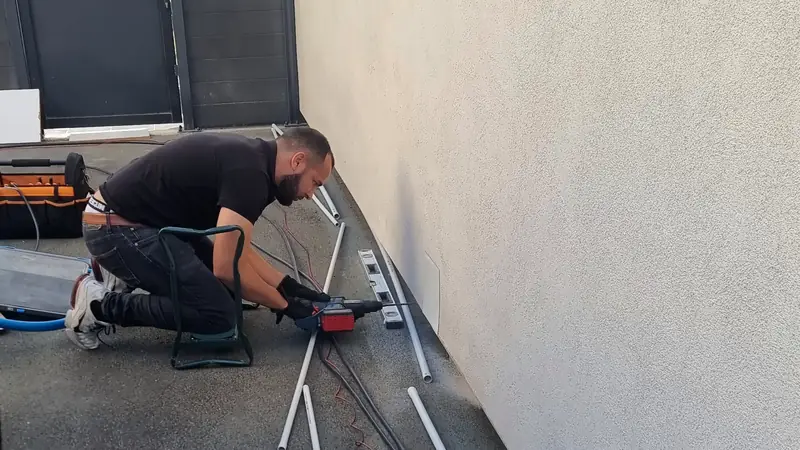 Le technicien pose les tubes gris du câble le long du mur d’une allée, jusqu’à la clôture du fond