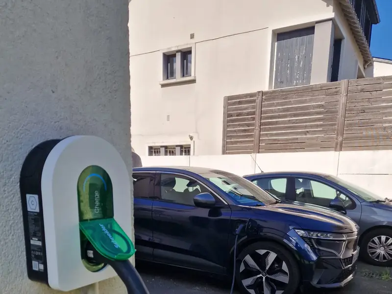 Borne Schneider Charge au clapet vert sur l’angle d’une maison, une voiture électrique bleu nuit branchée dans l’allée, en plein soleil