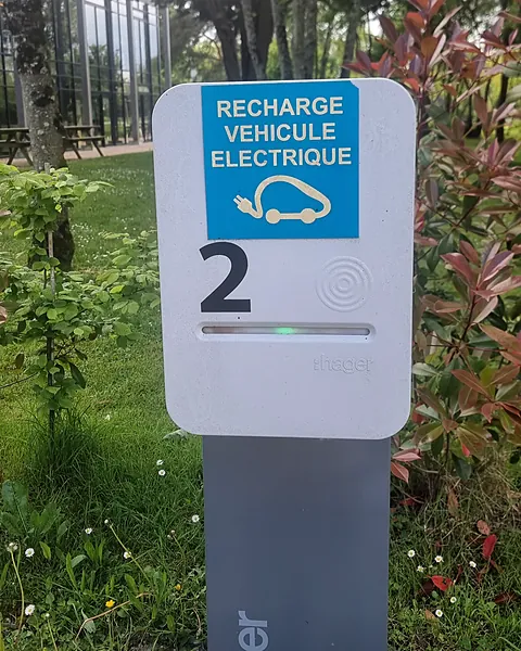 Borne de recharge Hager sur pied, refermée, voyant vert allumé, dans un espace vert : panneau « Recharge véhicule électrique » et numéro de place