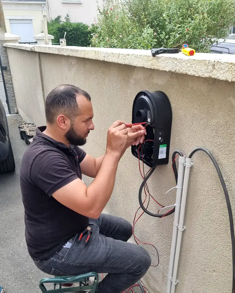 Dehors, devant un muret crépi surmonté d’une haie : un technicien d’Ave Green raccorde au tournevis isolé une borne de recharge Schneider Charge ouverte et fixée au mur ; des gaines grises et des câbles noirs descendent le long du muret.