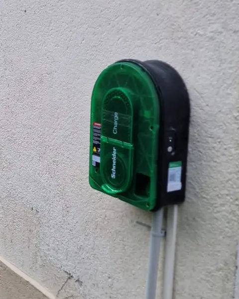 Borne Schneider Charge à façade verte translucide et coque noire sur un mur crépi clair ; deux tubes gris descendent de la borne et longent le soubassement.