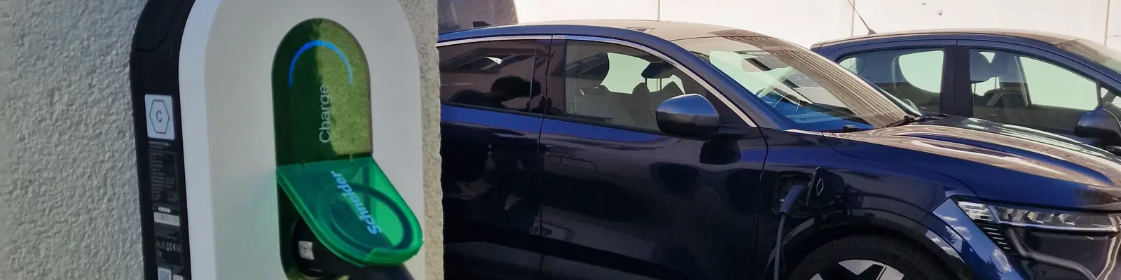 Borne de recharge Schneider Charge blanche à clapet vert, posée sur le mur crépi d’une maison, pistolet branché sur une voiture électrique bleu nuit en charge