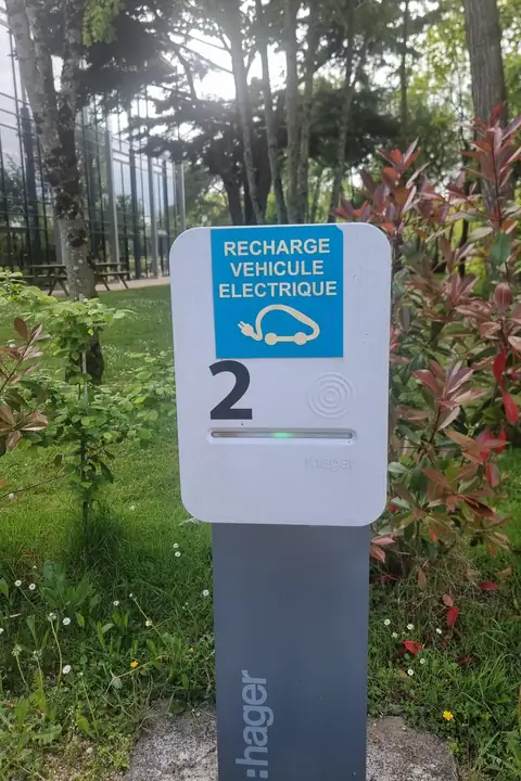 Borne de recharge sur pied numérotée 2, voyant vert allumé, devant une pelouse et un bâtiment vitré