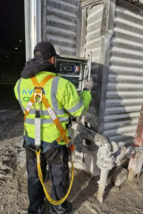 Électricien d’Ave Green de dos, en veste haute visibilité et harnais, devant une armoire électrique fixée sur un bardage