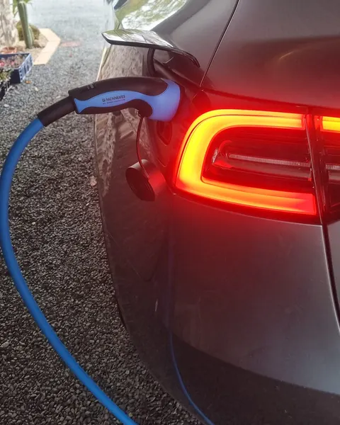 Voiture électrique grise en charge : pistolet de recharge bleu et noir branché dans la trappe, câble bleu posé sur le gravier, feu arrière allumé