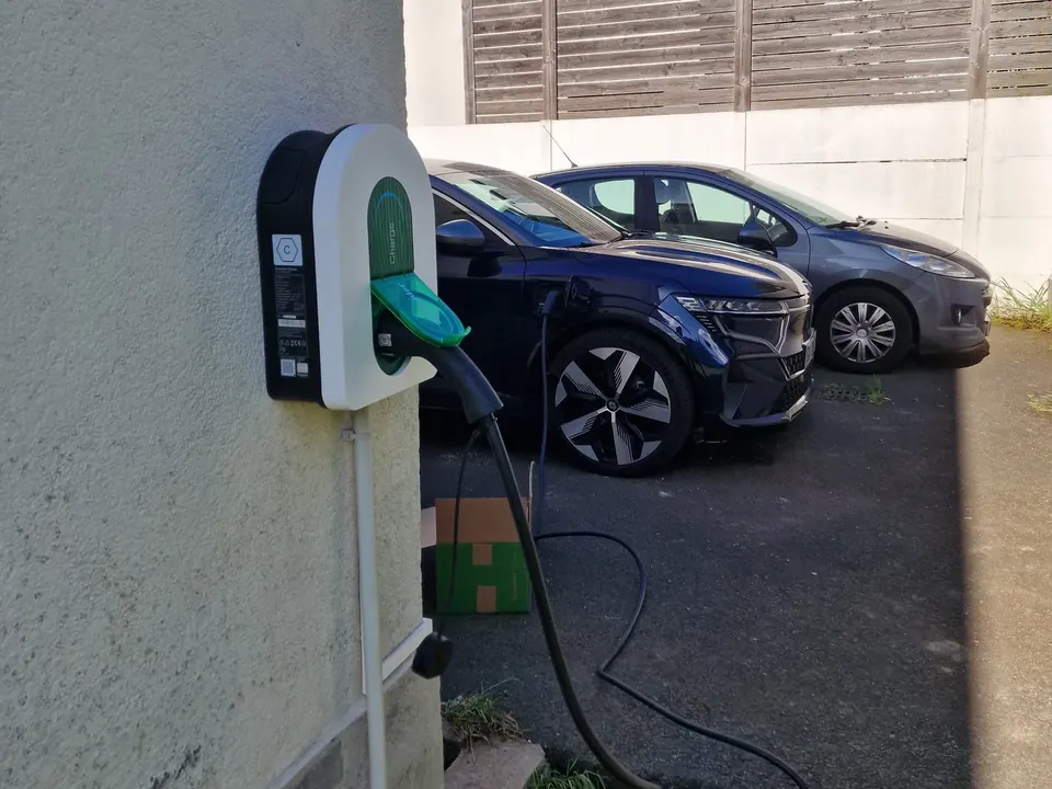 Borne murale blanche à trappe verte fixée à l’angle d’une maison, son câble noir branché à une voiture électrique bleu nuit garée dans la cour