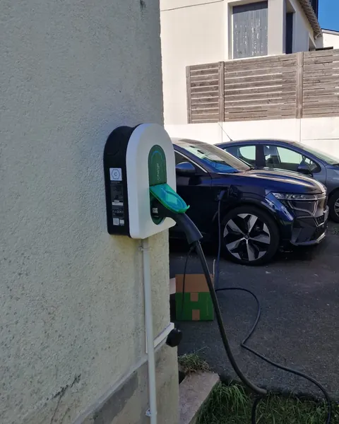 Borne murale blanche à trappe verte au coin d’une maison, câble branché à une voiture électrique bleu nuit dans la cour