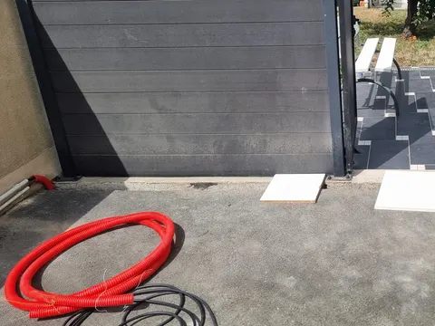 Couronne de gaine annelée rouge et câbles noirs posés sur le sol d’une cour, devant un portail