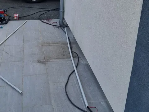 Tube gris et câble noir posés au pied d’une façade, le long d’une terrasse dallée, pendant la pose