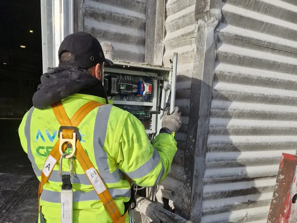 Électricien d’Ave Green vu de dos, en veste haute visibilité et harnais antichute, au travail sur une armoire électrique ouverte, fixée au bardage métallique d’un site industriel