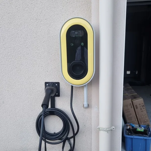 Une borne à cadre jaune fixée sur un mur clair, son câble noir rangé sur un crochet, à côté d’une descente d’eau