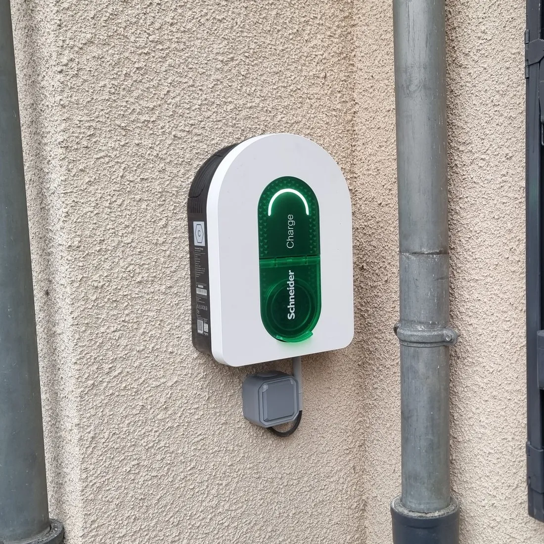 Une borne Schneider Charge blanche à vitre verte posée sur un mur crépi beige, entre deux descentes d’eau grises, à côté d’un volet en bois sombre