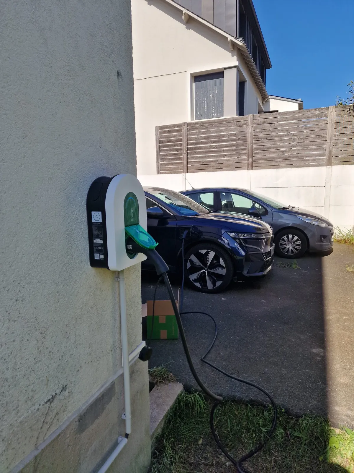 Une borne de recharge Schneider Charge posée sur le pignon d’une maison, branchée sur un SUV bleu nuit en charge, devant les maisons du quartier