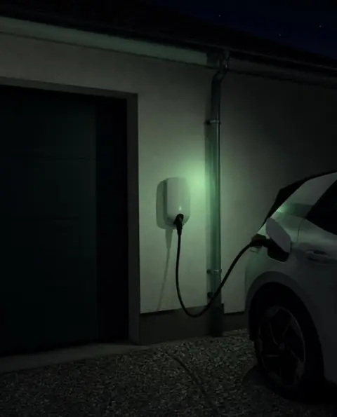 Borne murale blanche sans marque, la nuit, voyant vert allumé, câble branché sur une voiture blanche garée devant un garage