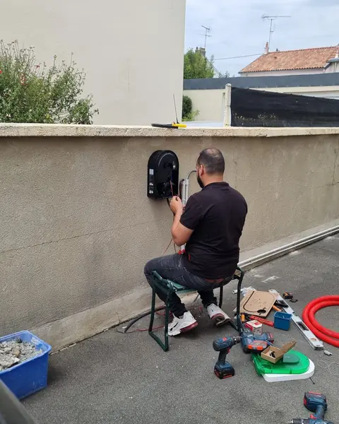 Électricien de dos, assis sur un tabouret, raccordant une borne murale noire sur un muret crépi, outils posés au sol