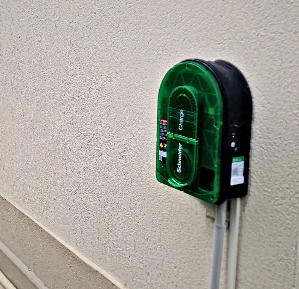 Borne de recharge Schneider Charge à façade verte posée dehors sur le mur enduit d’une maison ; deux tubes gris descendent de la borne vers le soubassement