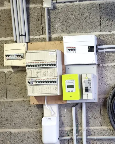 Tableau électrique d’une maison sur un mur en parpaings : tableau existant étiqueté, compteur Linky, disjoncteur de branchement et nouveau coffret blanc relié par des conduits gris