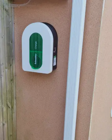 Borne Schneider Charge blanche à trappe verte fixée sur la façade enduite d’une maison, à côté d’une descente d’eau blanche