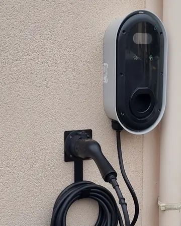 Borne murale ovale grise à façade noire vitrée, fermée, fixée sur le mur enduit beige d’une maison à côté du garage, câble noir enroulé sur son support et pistolet rangé, descente d’eau à droite