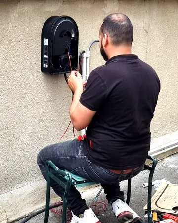 Le technicien d’Ave Green, de dos, assis sur un tabouret, raccorde une borne de recharge noire fixée sur un muret crépi, dehors