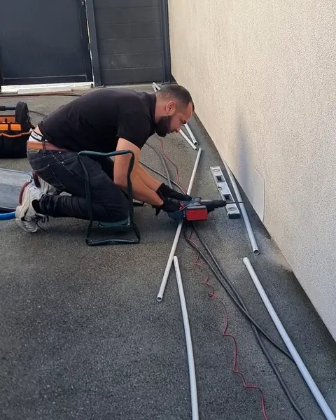 Technicien d’Ave Green à genoux au pied d’une façade claire, perceuse en main, tubes gris et câbles posés au sol le long du mur